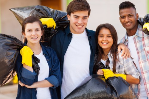 Image showing four young adults participating in a community rubbish removal activity outdoors, standing close together and smiling at the camera. They are dressed casually, with some wearing yellow rubber gloves and holding large black garbage bags filled with waste. The individuals appear to be volunteers or members of a local rubbish clearance group, possibly associated with houses or communal areas in Ware, as part of a domestic or neighbourhood cleanup initiative. The background features a blurry view of a residential or park area, indicating a suburban or mixed-use environment. The scene reflects a positive and environmentally conscious approach to rubbish disposal and house clearance, consistent with services offered by companies like House Clearance Ware in the local postcode vicinity. The lighting is natural, suggesting daytime conditions, and the overall mood conveys community involvement and environmental responsibility in waste management activities.