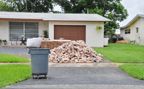 A pile of dismantled brickwork, consisting of various light brown and reddish bricks with rough and uneven surfaces, is situated on a paved driveway in front of a single-story house with a white exterior and brown garage door. The bricks are stacked loosely, spilling onto the concrete surface. To the left of the brick pile, a large grey wheeled rubbish bin with a closed lid is positioned on the edge of the driveway. The house's front features a large window with white frames, partially obscured by nearby greenery, including a small bush hanging from the wall. The surrounding area includes well-maintained green grass lawns on either side of the driveway, with a mature tree overhanging in the background, providing shade. The overall scene appears to be part of a house clearance or rubbish removal process, in a residential neighbourhood within Ware, as indicated by the context of the website houseclearanceware.co.uk.