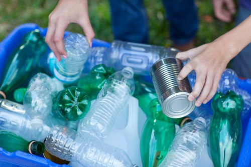 A pair of hands reaching into a blue plastic recycling bin filled with various discarded plastic bottles and containers, including clear and green-tinted bottles, some with caps and labels, placed outdoors on a grassy area possibly near a residential property. The person is handling a metal can and a transparent plastic bottle, indicating the process of sorting rubbish for collection or disposal. The background suggests a natural setting with grass visible around the bin. This scene exemplifies waste sorting efforts that professional rubbish removal services like House Clearance Ware might facilitate, particularly in residential areas of Ware postcodes. The image emphasizes environmental responsibility and proper waste separation practices in line with local rubbish clearance and recycling standards.