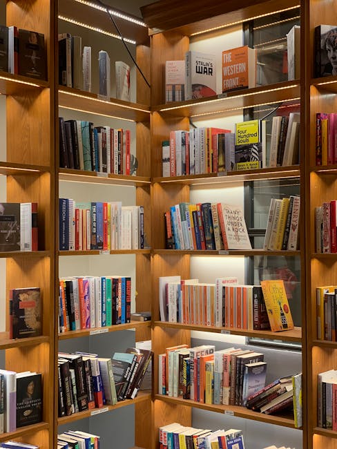 A tall wooden bookshelf filled with a diverse collection of hardcover and paperback books arranged horizontally and vertically on multiple shelves. The wood has a warm finish with visible grain textur