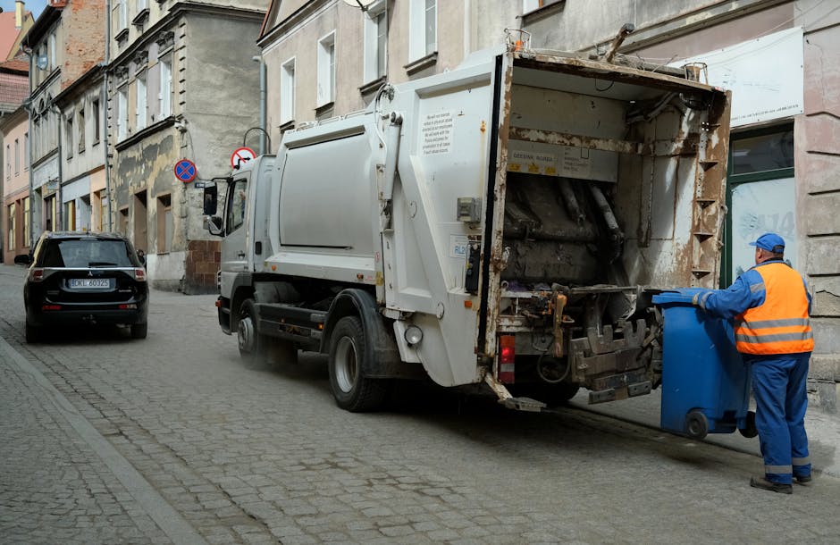 A narrow urban alleyway cluttered with scattered rubbish and debris, featuring a large black garbage bag in the foreground, partially torn and overflowing with waste. Behind it, various discarded item